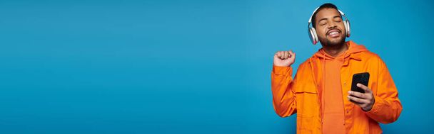 banner van lachende Afrikaans-Amerikaanse man in oranje outfit en hoofdtelefoon dansen met smartphone - Foto, afbeelding