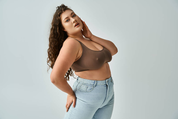 attractive plus size girl in brown bra and blue jeans posing and touching with hand to face - Photo, Image