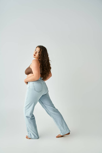 attractive curvy young woman in brown bra and blue jeans posing against grey background - Photo, Image