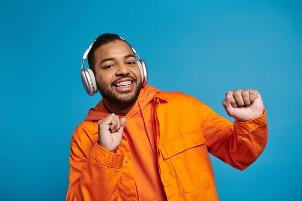 cheerful african american man in headphones dancing and looking to camera on blue background - Fotó, kép