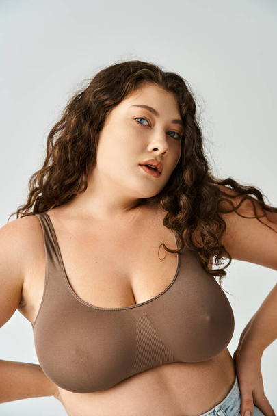 attractive plus size woman in brown bra and blue jeans posing with hands on waist on grey background - Photo, Image