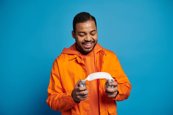 усміхнений афроамериканський чоловік в помаранчевому вбранні тримає навушники на синьому фоні - Фото, зображення