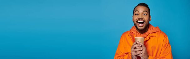 estandarte de encantador afroamericano joven en traje naranja sosteniendo con las manos taza de papel - Foto, imagen