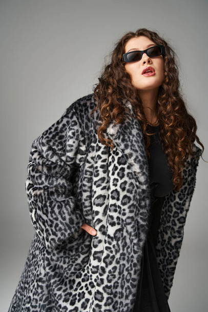 Gri arkaplanda poz veren leopar kürklü ve güneş gözlüklü şık ve kilolu genç bir kadın. - Fotoğraf, Görsel