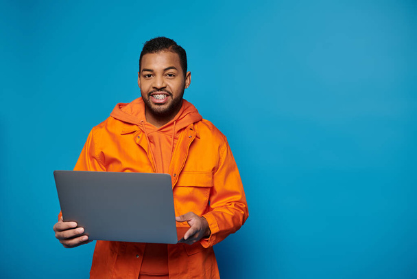 Turuncu kıyafetli neşeli Afro-Amerikan adam elinde dizüstü bilgisayarla kameraya bakıyor. - Fotoğraf, Görsel