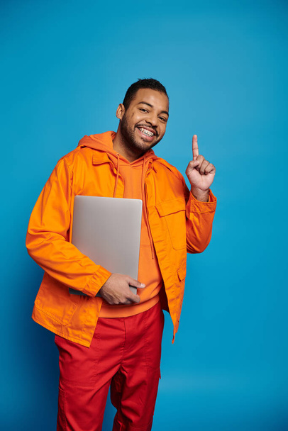 gelukkig afrikaans amerikaans man in oranje outfit poseren met laptop en putting vinger naar boven - Foto, afbeelding