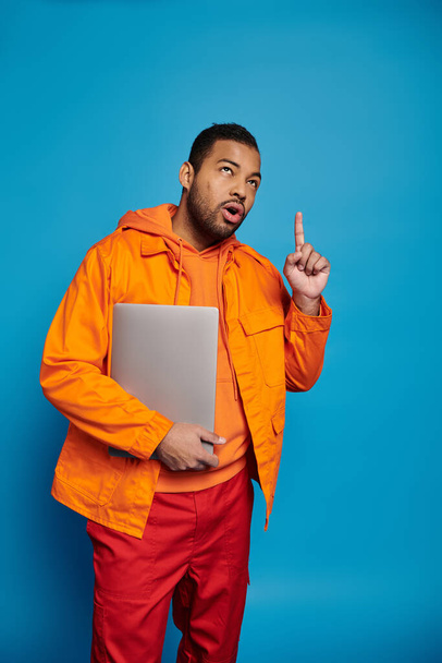 hombre afroamericano reflexivo en traje naranja posando con el ordenador portátil y poniendo el dedo para arriba - Foto, imagen