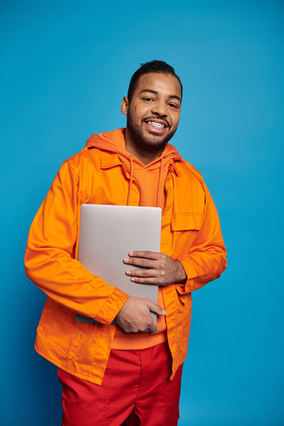 Gülümseyen Afro-Amerikalı turuncu elbiseli genç adam mavi arka planda dizüstü bilgisayarla poz veriyor. - Fotoğraf, Görsel