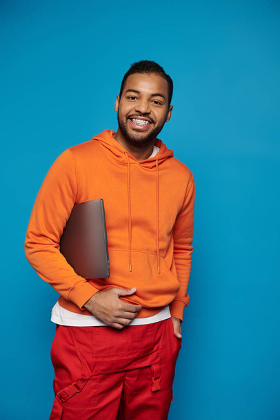 attractive african american guy in vibrant outfit posing with laptop under arm on blue background - Foto, Imagen
