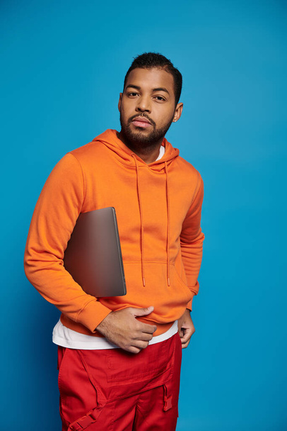 charismatic african american man in vibrant outfit posing with laptop under arm on blue background - Foto, Imagem