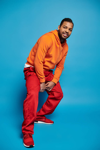 handsome african american man in vibrant outfit leaning and crouching on blue background - Foto, imagen