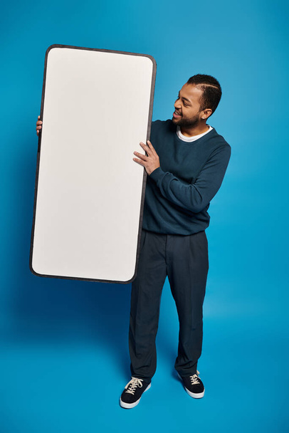 souriant homme afro-américain tenant et regardant la maquette de smartphone sur fond bleu - Photo, image