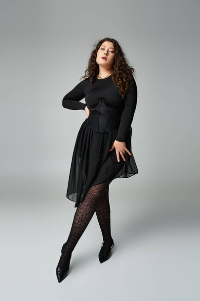 verführerische kurvige Frau im schwarzen Outfit, die das Bein mit der Hand nach vorne auf die Taille auf grauem Hintergrund legt - Foto, Bild