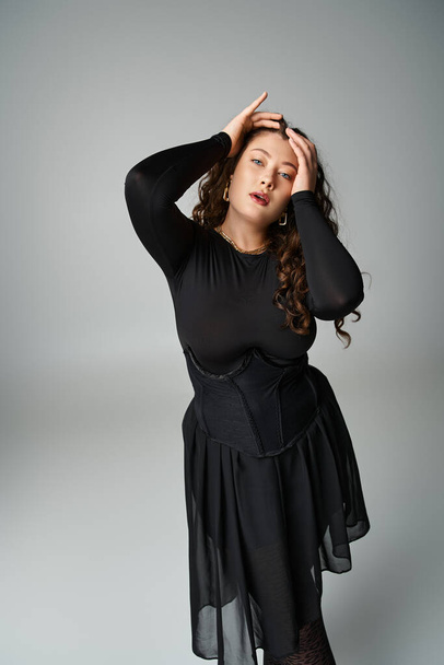encantadora jovem curvilínea em roupa elegante preto de mãos dadas na cabeça no fundo cinza - Foto, Imagem