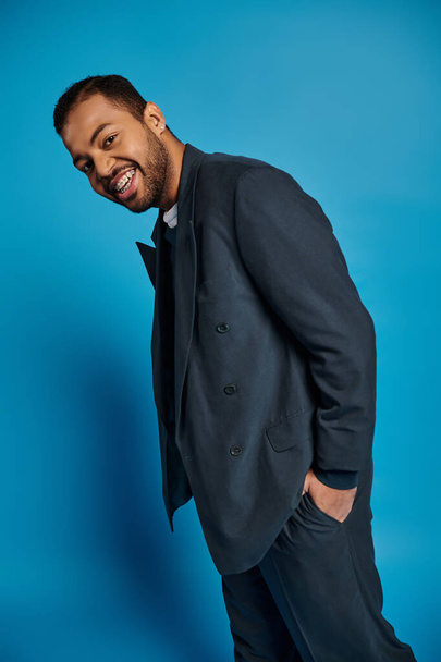 fröhlicher afrikanisch-amerikanischer junger Mann im dunkelblauen Outfit, der mit der Hand in der Tasche zur Seite steht - Foto, Bild