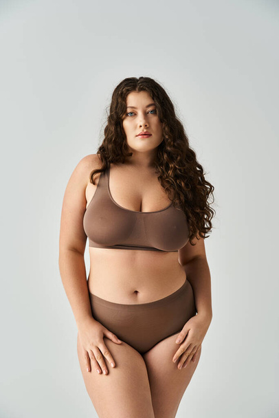 attractive plus size woman in brown lingerie with curly hair and blue eyes posing on grey background - Photo, Image