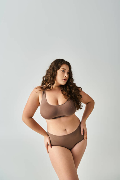 charming curvy girl in brown underwear with curly hair posing hands on hips and looking to side - Photo, Image
