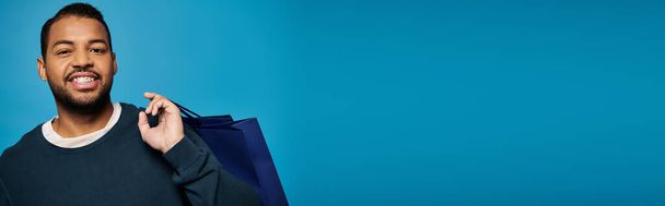 banner of happy african american young man with shopping bag over shoulder on blue background - Photo, Image