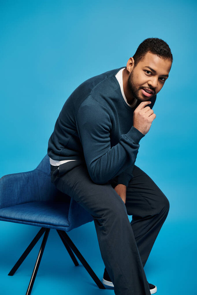 encantador hombre afroamericano sentado en el respaldo de la silla y tocando la barbilla sobre fondo azul - Foto, Imagen