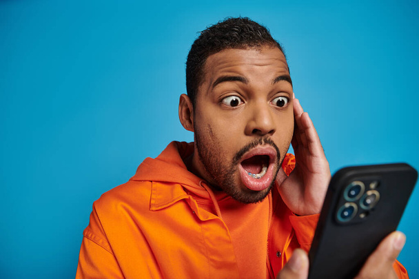 megdöbbent afro-amerikai férfi görgetés szociális média okostelefon kék háttér - Fotó, kép