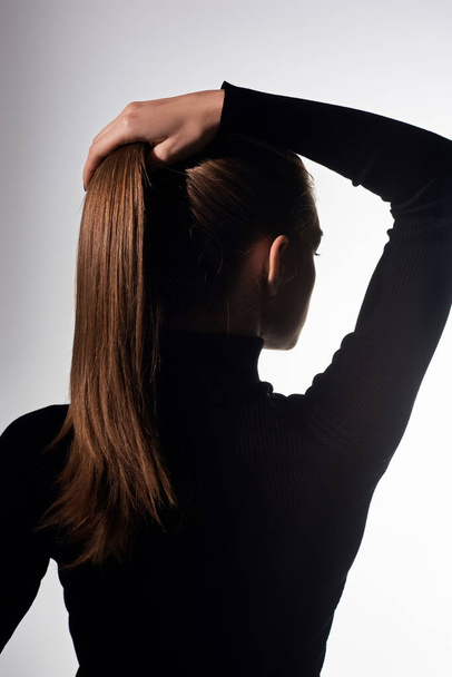 Eine junge schöne Frau mit langen Haaren hält sich die Haare, während sie einen schwarzen Rollkragen trägt. - Foto, Bild