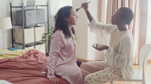 Mittlere lange Aufnahme eines schwarzen Mädchens, das ihren Freund schminkt, der auf dem Bett sitzt, während er sich auf eine Party zu Hause vorbereitet - Filmmaterial, Video