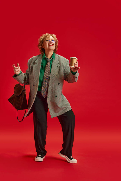 Friday mood. Overjoyed senior lady, dressed elegant fashion office outfit and dancing against red studio background. Concept of business, fashionable older adults, active seniors in modern life - Foto, imagen