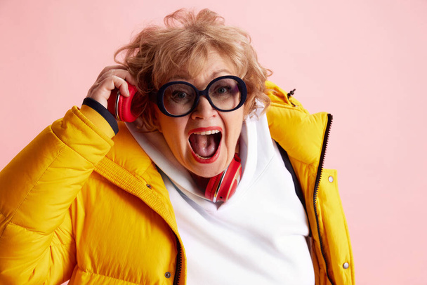 Emocionální starší žena poslouchající hudbu ve sluchátkách a zpívající ve žluté bundě. Koncept posílení postavení starších dospělých, aktivních seniorů v moderním životě, seniorů využívajících technologie. Reklama - Fotografie, Obrázek
