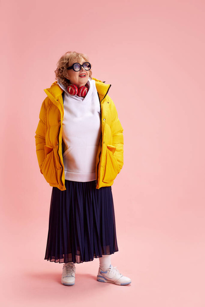 Full length portrait of elderly fashionista in warm yellow jacket, skirt, white sneakers looks upbeat against pink background. Concept of active seniors in modern life, seniors using technology. - Photo, image