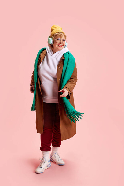 Full length portrait of beautiful stylish dressed mature woman listening music in headphones and dancing against pink background. Concept of active seniors in modern life, seniors using technology. - Zdjęcie, obraz