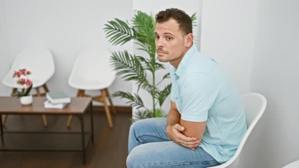 Hombre hispano guapo con los brazos cruzados sentado en un interior moderno con plantas y sillas blancas. - Metraje, vídeo