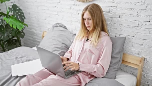 Una donna bionda in pigiama rosa che lavora su un computer portatile nella sua camera da letto, con un'espressione stanca che si strofina gli occhi. - Filmati, video
