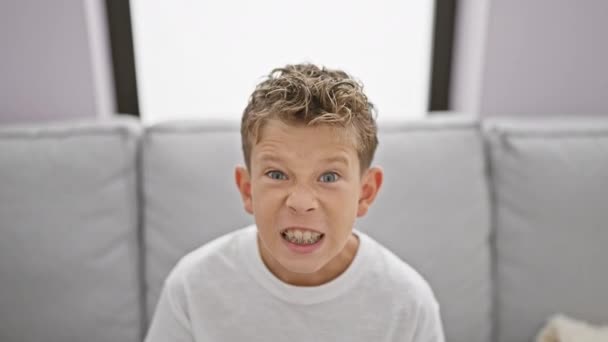 Imádnivaló szőke fiú ül a kanapén otthon, kifejezve hangos frusztráció és harag - Felvétel, videó