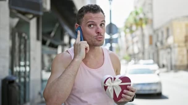 Komea mies puhuu puhelimessa ja pitää lahjapakkausta aurinkoisella kadulla. - Materiaali, video