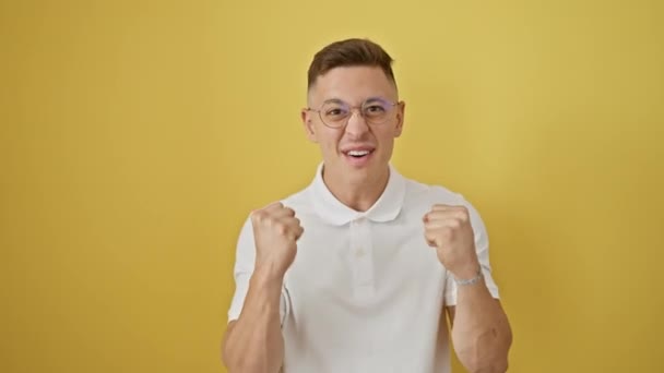 A diadalmas fiatal spanyol szemüveges férfi örömteli győzelmet ünnepel, elszigetelt sárga háttérrel szemben, magabiztosságot és boldogságot árasztva.. - Felvétel, videó