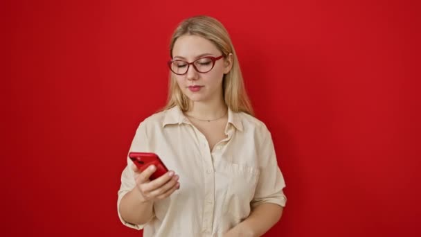 Radosna młoda blondynka celująca kciukiem w bok, z otwartymi ustami uśmiech nad czerwoną ścianą, entuzjastycznie pokazująca coś za pomocą smartfona - Materiał filmowy, wideo