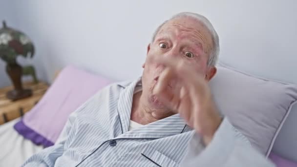 Sorprendido hombre mayor en pijama, sentado en la cama de su dormitorio, mirando a través de los dedos sorprendidos con expresión incrédula. gesto divertido que significa ok! - Metraje, vídeo