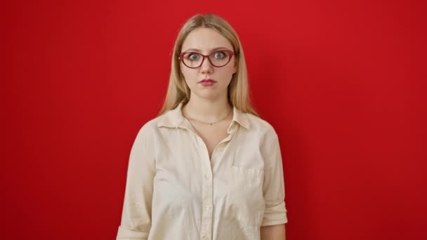 Νεαρή ξανθιά γυναίκα, σκεπτική και σαρκαστική, στέκεται σε δυσπιστία πάνω από ένα κόκκινο απομονωμένο φόντο, με μια κυνική ματιά και ανοιχτό το στόμα έκφραση του σοκ - Πλάνα, βίντεο