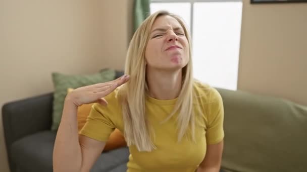 Furious young blonde woman in t-shirt threatens violence at home, cutting air at her throat with hand like a knife in angry gesture - Footage, Video