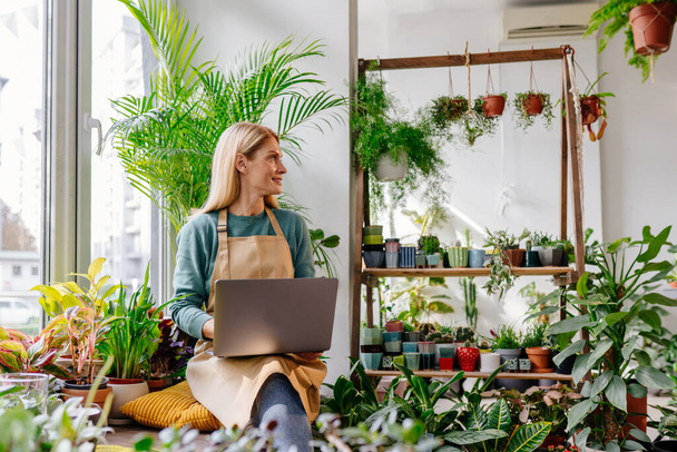 Hymyilevä keski-ikä vaalea nainen liiketoiminnan nainen kukkakauppa käyttäen digitaalista kannettavaa katselee sivuun vehreys studio sisustus. - Valokuva, kuva