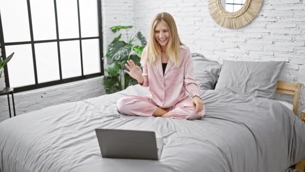 Pembe pijamalı genç bir kadın modern bir yatak odasında dizüstü bilgisayarla yatağa uzanıyor. - Video, Çekim