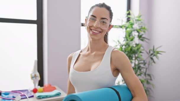 Mulher hispânica jovem confiante ostentando um belo sorriso e segurando seu tapete de ioga na clínica de reabilitação - Filmagem, Vídeo