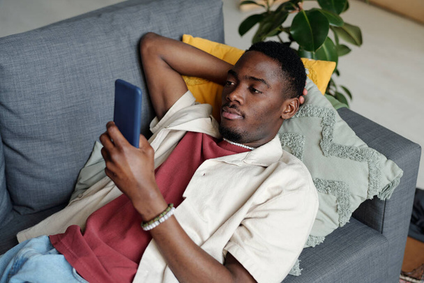 Jongeman in casualwear ontspannen op de bank met kussens en kijken naar het scherm van de smartphone tijdens het kijken naar online video of film - Foto, afbeelding
