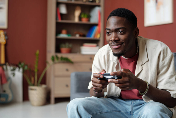 Sonriente chico afroamericano pulsando botones en los controladores y mirando a la pantalla de la computadora o televisor mientras juega videojuego - Foto, imagen