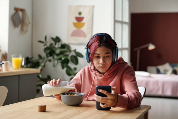 Cute Asian female teenager in headphones pouring fresh milk into bowl with cornflakes and looking at screen of smartphone in her hand - Photo, Image