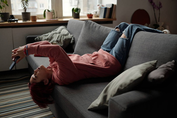 Bastante adolescente en ropa de casualwear acostado en un cómodo sofá en la sala de estar y mirando la pantalla del teléfono inteligente mientras ve video en línea - Foto, Imagen