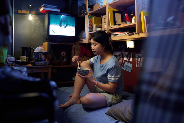 Jonge Aziatische vrouw met eetstokjes in kom van Chinees eten zitten op bed in klein appartement, tv kijken en lunchen - Foto, afbeelding
