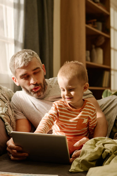Rijpere vader en baby zoon kijken naar tablet scherm terwijl ontspannen op bed of bank in de voorkant van de camera en samen kijken naar online video - Foto, afbeelding