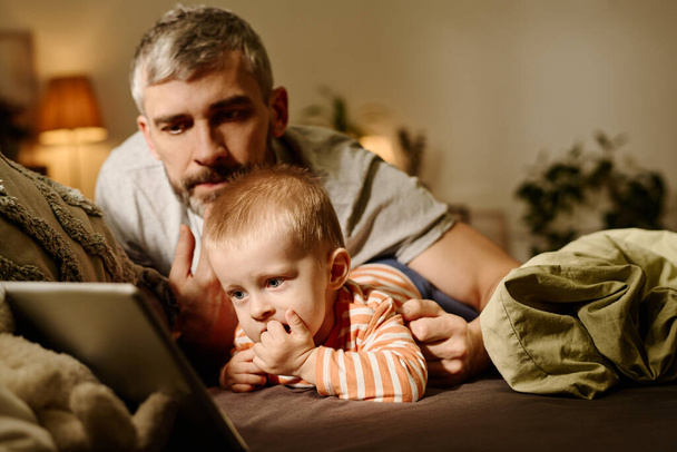 Starší otec ležící na posteli vedle sladké dítě syna, zatímco oba při pohledu na obrazovce tabletu a sledování online karikatury ve večerních hodinách - Fotografie, Obrázek