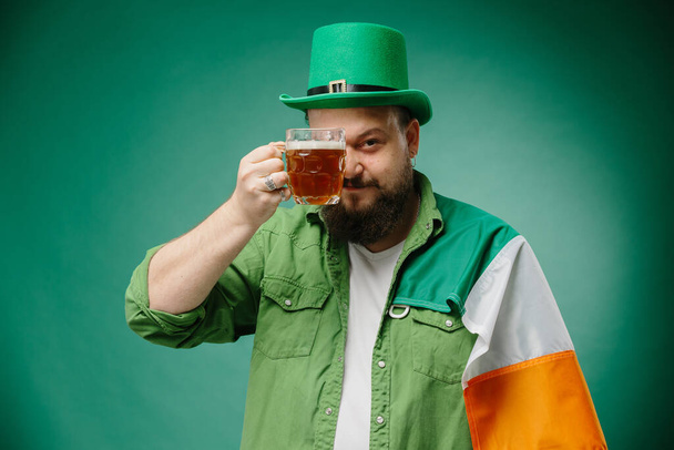 Счастливый бородатый мужчина со стаканом пива и ирландским флагом на темно-зеленом фоне. День Святого Патрика. - Фото, изображение